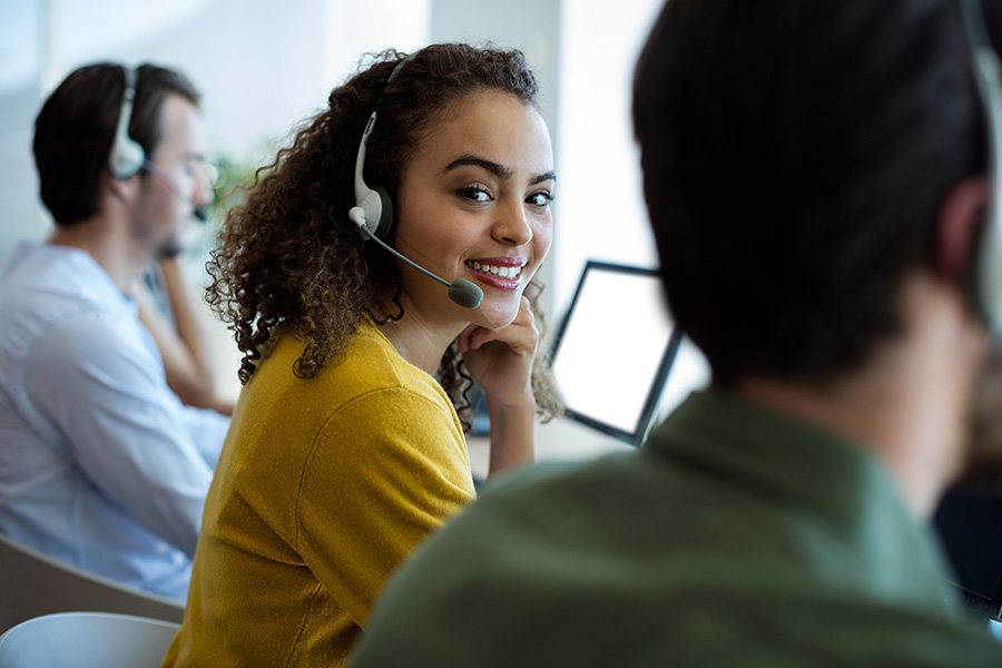 Billing Accounting Questions - Portrait of a Smiling Young Woman Wearing a Headset While Working in a Support Center Office Next to Her Colleagues